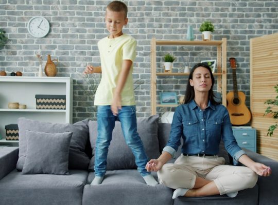 Happy kid boy is dancing on sofa while young mother is meditating in lotus position at home sitting on couch with closed eyes. Childhood and parenthood concept.