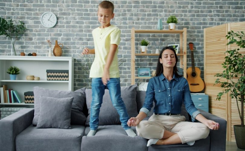 Happy kid boy is dancing on sofa while young mother is meditating in lotus position at home sitting on couch with closed eyes. Childhood and parenthood concept.